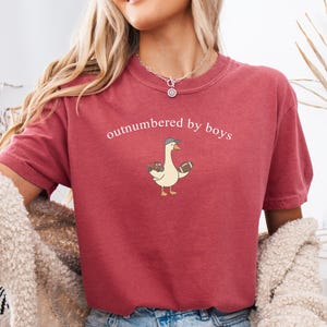 May include: A maroon t-shirt with the phrase "outnumbered by boys" printed across the chest. The shirt features a cartoon goose wearing a baseball cap and holding a football and toy trucks. The shirt has short sleeves and a crew neck.
