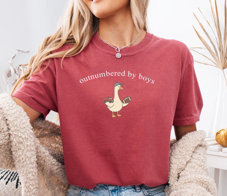 May include: A maroon t-shirt with the phrase "outnumbered by boys" printed across the chest. The shirt features a cartoon goose wearing a baseball cap and holding a football and toy trucks. The shirt has short sleeves and a crew neck.