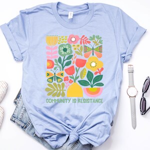 Op de afbeelding: Lichtblauw T-shirt met een kleurrijk bloemenmotief en de tekst "Community is Resistance".