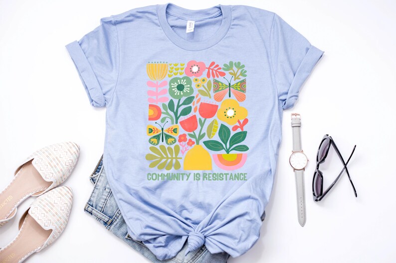 Op de afbeelding: Lichtblauw T-shirt met een kleurrijk bloemenmotief en de tekst "Community is Resistance".