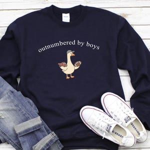 May include: Navy blue sweatshirt with the text "outnumbered by boys" above a cartoon goose wearing a cap and holding a football and toy cars. The sweatshirt is paired with blue jeans and white sneakers.
