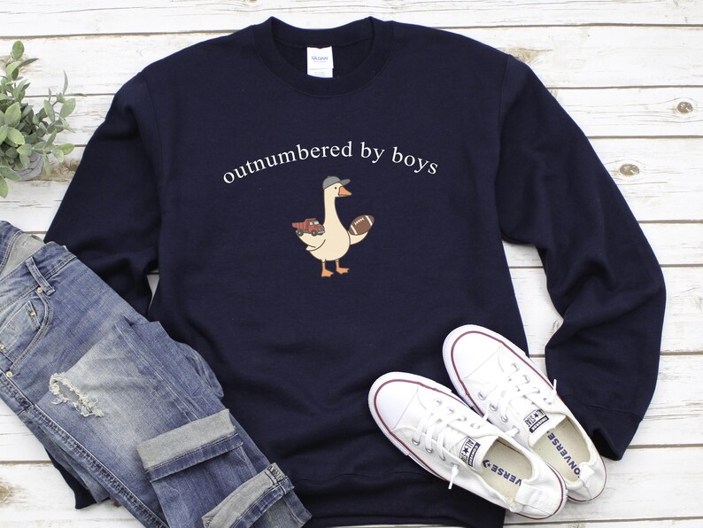 May include: Navy blue sweatshirt with the text "outnumbered by boys" above a cartoon goose wearing a cap and holding a football and toy cars. The sweatshirt is paired with blue jeans and white sneakers.