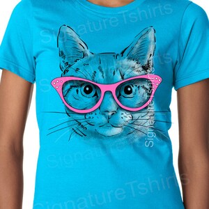 May include: A turquoise blue t-shirt with a graphic of a cat wearing pink glasses. The cat's eyes are blue and the glasses have rhinestones.