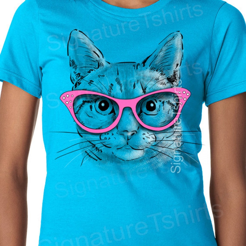 May include: A turquoise blue t-shirt with a graphic of a cat wearing pink glasses. The cat's eyes are blue and the glasses have rhinestones.