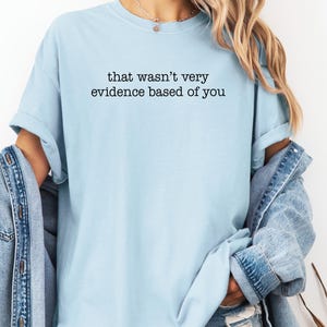 May include: Light blue t-shirt with the text "that wasn't very evidence based of you" in black. The shirt has short sleeves and a relaxed fit. A denim jacket is draped over the shoulders.