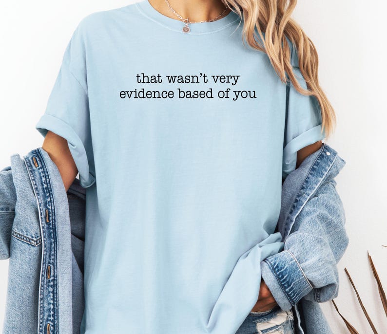 May include: Light blue t-shirt with the text "that wasn't very evidence based of you" in black. The shirt has short sleeves and a relaxed fit. A denim jacket is draped over the shoulders.