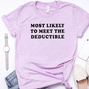 May include: A light purple t-shirt with the text "MOST LIKELY TO MEET THE DEDUCTIBLE" in black. The shirt is styled with a gray watch, a smartphone, a pair of cream-colored flats, and a pair of blue jeans.