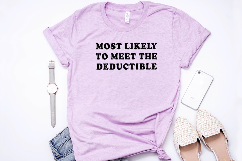 May include: A light purple t-shirt with the text "MOST LIKELY TO MEET THE DEDUCTIBLE" in black. The shirt is styled with a gray watch, a smartphone, a pair of cream-colored flats, and a pair of blue jeans.