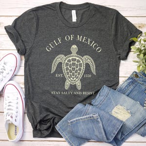 Puede incluir: Camiseta gris oscuro con un gr&aacute;fico blanco de tortuga marina y el texto "GULF OF MEXICO EST. 1550 STAY SALTY AND RESIST." La imagen tambi&eacute;n muestra vaqueros azules y zapatillas de lona blancas.