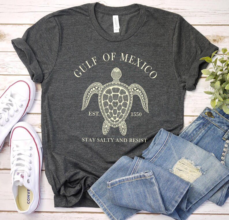 Puede incluir: Camiseta gris oscuro con un gr&aacute;fico blanco de tortuga marina y el texto "GULF OF MEXICO EST. 1550 STAY SALTY AND RESIST." La imagen tambi&eacute;n muestra vaqueros azules y zapatillas de lona blancas.