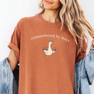 May include: A terracotta-coloured t-shirt featuring the text "outnumbered by boys" above a cartoon goose wearing a baseball cap and holding a football and a toy truck. The shirt has short sleeves and a relaxed fit.