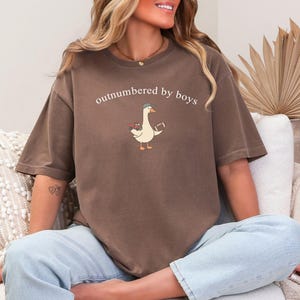 May include: A brown t-shirt featuring the text "outnumbered by boys" and a cartoon goose wearing a hat and holding a football. The goose also carries flowers. The shirt is worn with light blue jeans.