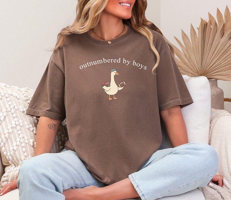 May include: A brown t-shirt featuring the text "outnumbered by boys" and a cartoon goose wearing a hat and holding a football. The goose also carries flowers. The shirt is worn with light blue jeans.