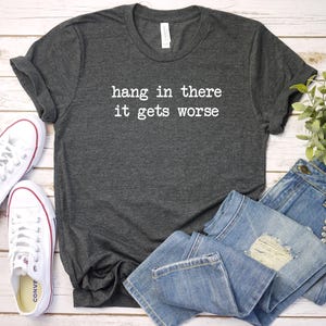 May include: Dark gray t-shirt with the text "hang in there it gets worse" in white. The shirt is paired with light blue denim jeans and white canvas sneakers.