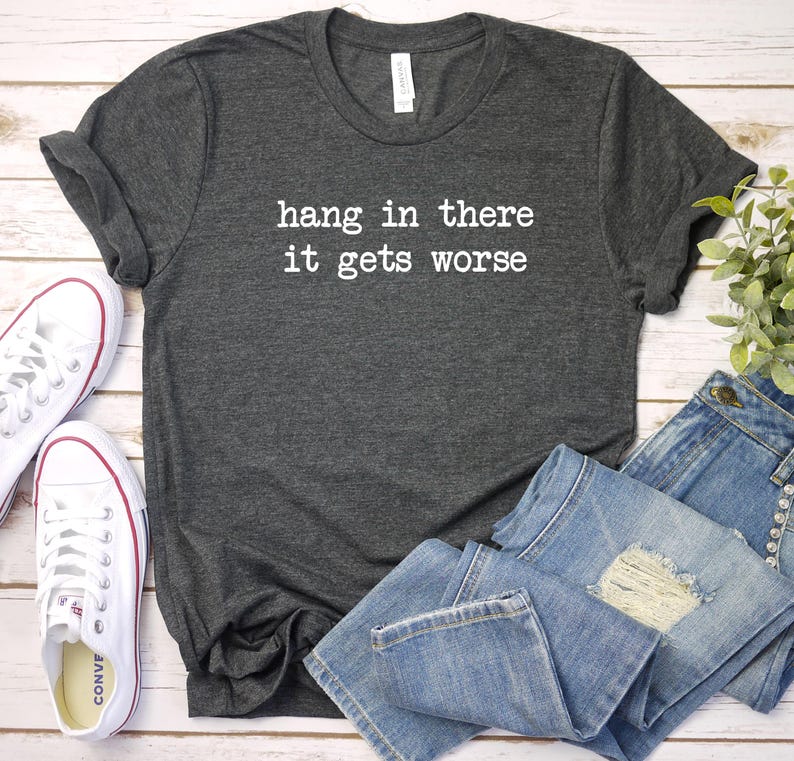 May include: Dark gray t-shirt with the text "hang in there it gets worse" in white. The shirt is paired with light blue denim jeans and white canvas sneakers.