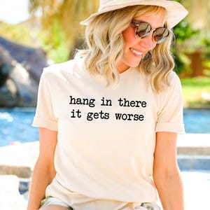 May include: Cream-colored t-shirt with the text "hang in there it gets worse" in black. The shirt is worn by a person wearing sunglasses and a hat. The background is blurred, suggesting an outdoor setting.