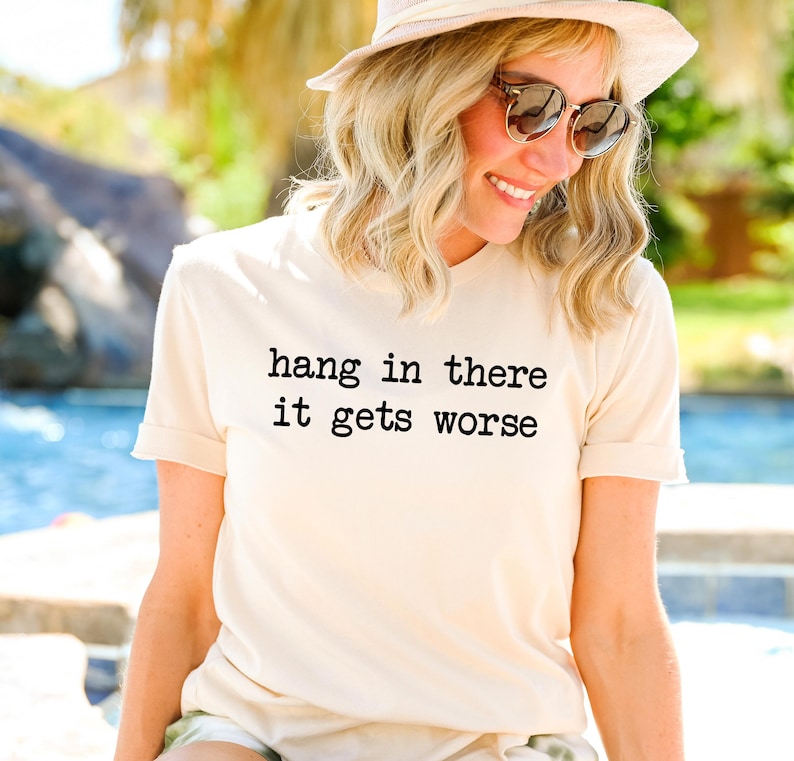 May include: Cream-colored t-shirt with the text "hang in there it gets worse" in black. The shirt is worn by a person wearing sunglasses and a hat. The background is blurred, suggesting an outdoor setting.