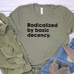 May include: An olive green t-shirt with the text "Radicalized by basic decency." is displayed with a matching jacket, blue jeans, and gray ankle boots. The items are arranged on a light-colored surface.