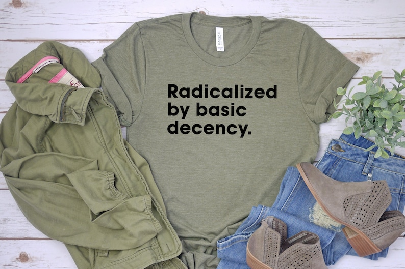 May include: An olive green t-shirt with the text "Radicalized by basic decency." is displayed with a matching jacket, blue jeans, and gray ankle boots. The items are arranged on a light-colored surface.