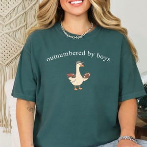May include: Teal t-shirt with the white text "outnumbered by boys". The shirt features a cartoon goose wearing a hat and holding a football and a red truck. The t-shirt is a casual, unisex style.