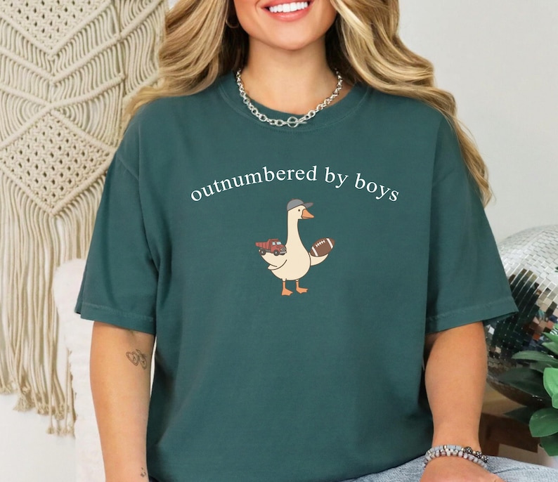 May include: Teal t-shirt with the white text "outnumbered by boys". The shirt features a cartoon goose wearing a hat and holding a football and a red truck. The t-shirt is a casual, unisex style.