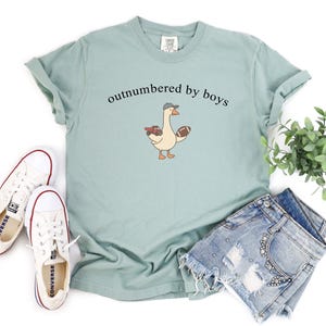 May include: A pale green t-shirt featuring the text "outnumbered by boys" above a cartoon goose wearing a cap and holding a rugby ball and a toy truck. White trainers and denim shorts are also in the image.