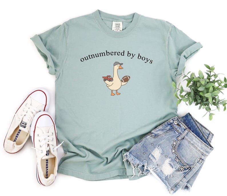 May include: A pale green t-shirt featuring the text "outnumbered by boys" above a cartoon goose wearing a cap and holding a rugby ball and a toy truck. White trainers and denim shorts are also in the image.