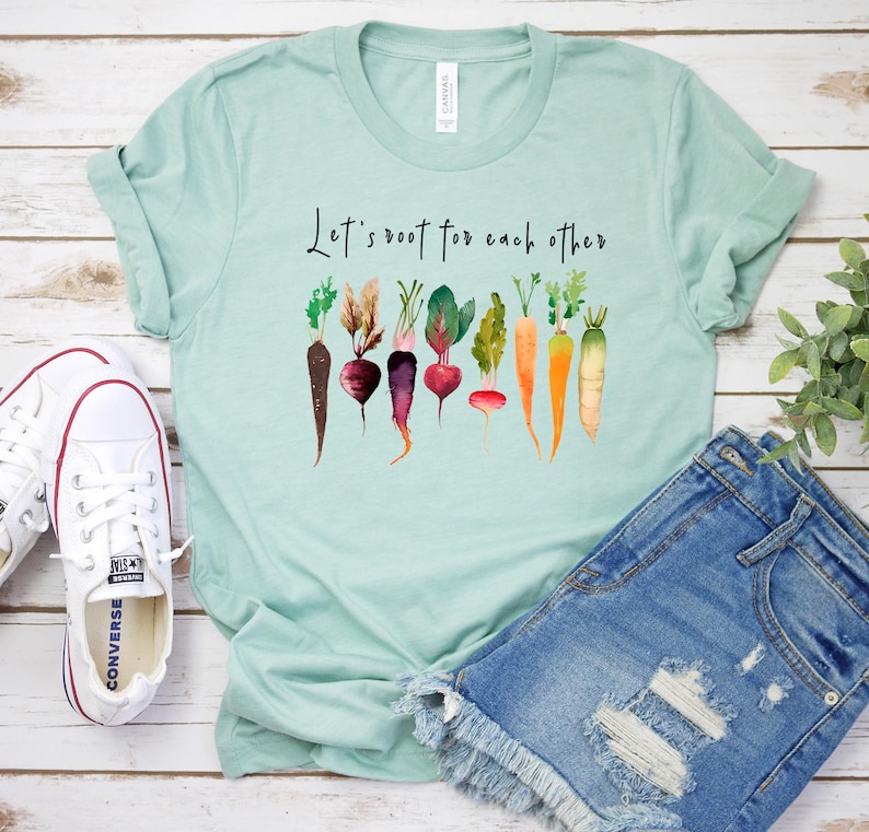 May include: A light blue t-shirt with the text "Let's root for each other" printed on the front. The shirt features a watercolor design of various vegetables, including carrots, beets, and radishes.