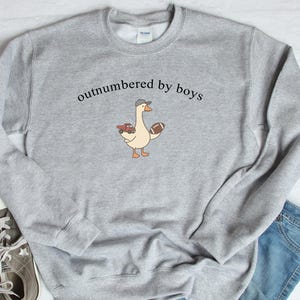 May include: A light gray sweatshirt with the text "outnumbered by boys" above a cartoon goose wearing a hat and holding a toy truck and football. The sweatshirt is paired with jeans and sneakers.