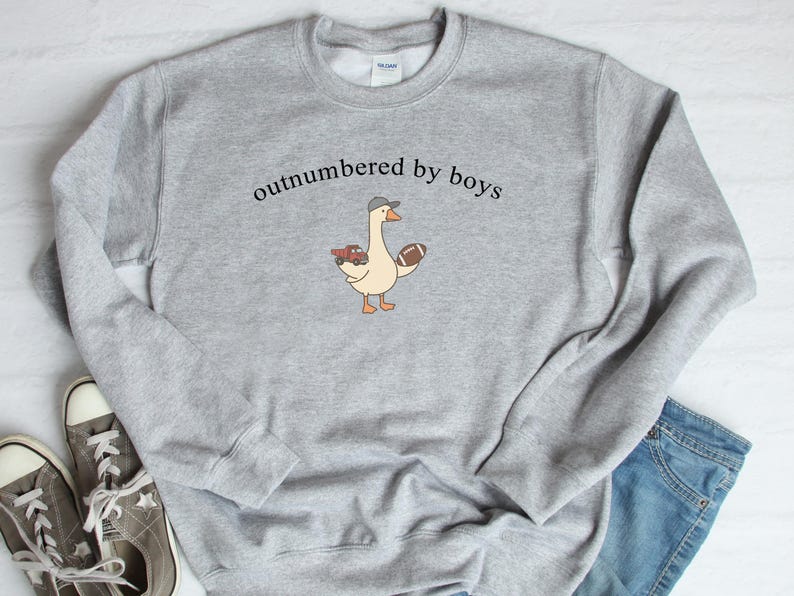 May include: A light gray sweatshirt with the text "outnumbered by boys" above a cartoon goose wearing a hat and holding a toy truck and football. The sweatshirt is paired with jeans and sneakers.