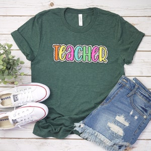 Puede incluir: Una camiseta verde con la palabra "teacher" escrita en letras de colores con un patr&oacute;n de lunares blancos.