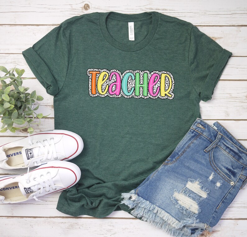 Puede incluir: Una camiseta verde con la palabra "teacher" escrita en letras de colores con un patr&oacute;n de lunares blancos.