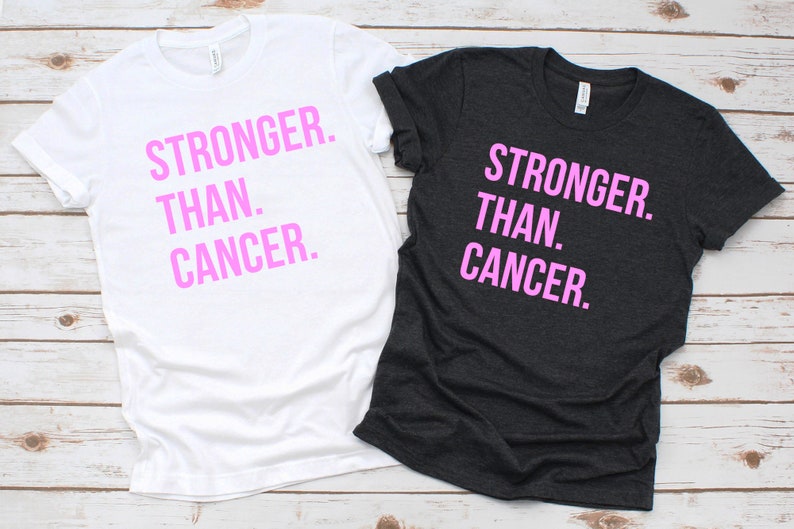 May include: Two t-shirts with the text "Stronger Than Cancer" printed in pink on the front. One t-shirt is white and the other is dark grey.