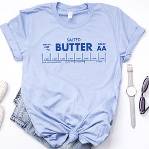 May include: Light blue t-shirt with a graphic of a stick of butter labeled "Salted Butter Grade AA" with a measurement guide in tablespoons and cups. The shirt is knotted at the bottom.