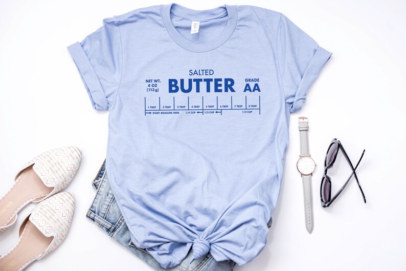 May include: Light blue t-shirt with a graphic of a stick of butter labeled "Salted Butter Grade AA" with a measurement guide in tablespoons and cups. The shirt is knotted at the bottom.