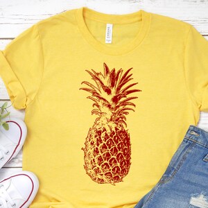 Pode incluir: Uma camiseta amarela com um gr&aacute;fico de abacaxi vermelho. O abacaxi &eacute; uma ilustra&ccedil;&atilde;o de estilo vintage.
