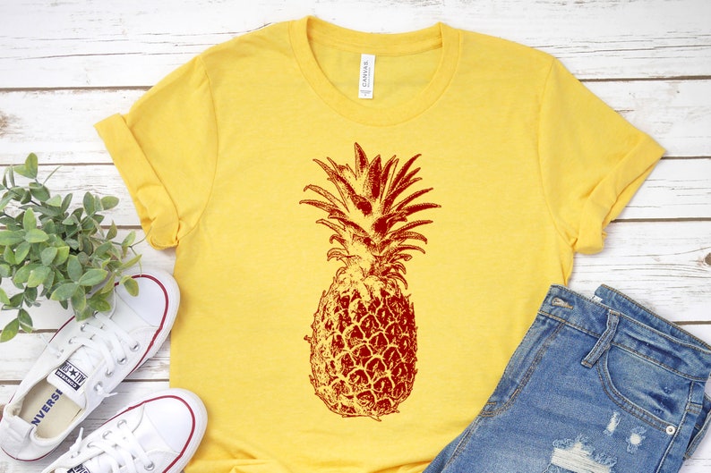 Pode incluir: Uma camiseta amarela com um gr&aacute;fico de abacaxi vermelho. O abacaxi &eacute; uma ilustra&ccedil;&atilde;o de estilo vintage.