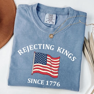May include: A blue t-shirt with the words "REJECTING KINGS" above an American flag graphic and the text "SINCE 1776" below. A gold necklace with a pendant rests on the shirt.