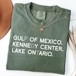 May include: An olive green crew neck t-shirt with white text that reads GULF OF MEXICO. KENNEDY CENTER. LAKE ONTARIO. The shirt features a Comfort Colors brand label at the inner neckline and is paired with a delicate gold-toned pendant necklace.