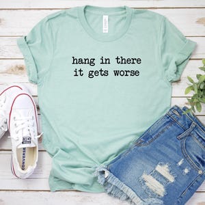 May include: A light green t-shirt with the text "hang in there it gets worse" in black. The shirt is paired with denim shorts and white sneakers with red accents. The items are arranged on a light wood surface.
