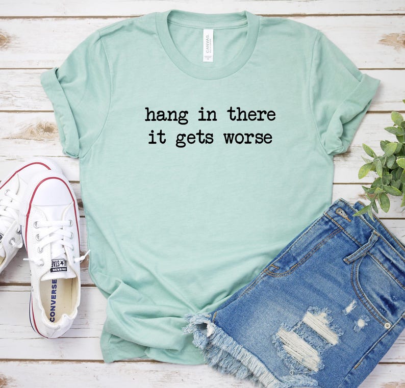 May include: A light green t-shirt with the text "hang in there it gets worse" in black. The shirt is paired with denim shorts and white sneakers with red accents. The items are arranged on a light wood surface.