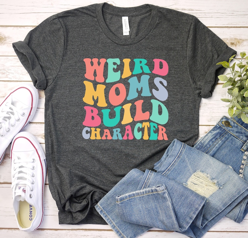 Pode incluir: Uma camiseta cinza escura com o texto "Weird Moms Build Character" em cores do arco-&iacute;ris. O texto est&aacute; em um estilo de fonte retr&ocirc;.