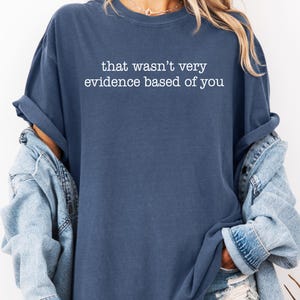 May include: A navy blue long-sleeve shirt with the text "that wasn't very evidence based of you" in white. The shirt is a relaxed fit and has rolled-up sleeves. A light blue denim jacket is worn over the shirt.