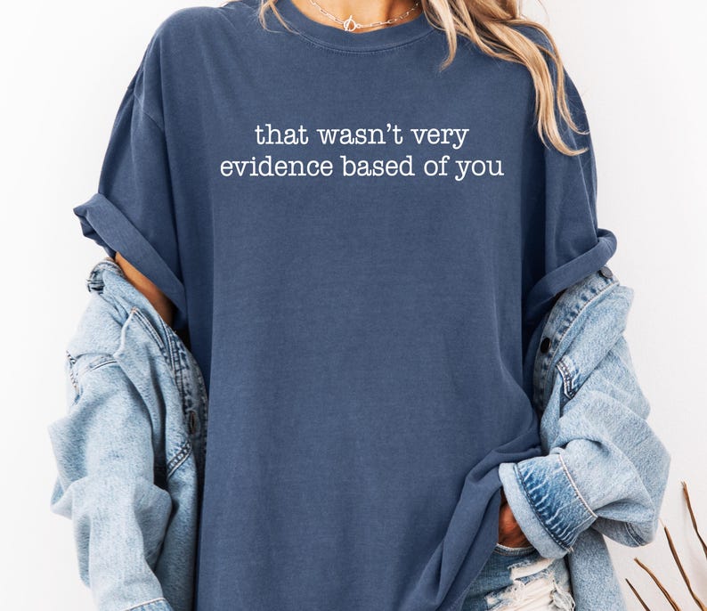 May include: A navy blue long-sleeve shirt with the text "that wasn't very evidence based of you" in white. The shirt is a relaxed fit and has rolled-up sleeves. A light blue denim jacket is worn over the shirt.