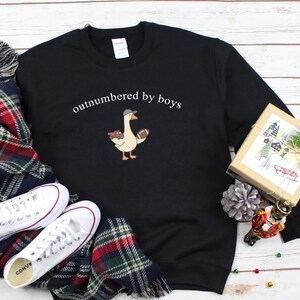 May include: Black sweatshirt with the text "outnumbered by boys" above a cartoon goose wearing a baseball cap and holding a football. White sneakers and a plaid scarf are next to the sweatshirt. A wrapped gift box and holiday decorations are also in the image.
