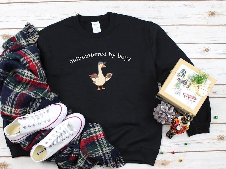 May include: Black sweatshirt with the text "outnumbered by boys" above a cartoon goose wearing a baseball cap and holding a football. White sneakers and a plaid scarf are next to the sweatshirt. A wrapped gift box and holiday decorations are also in the image.