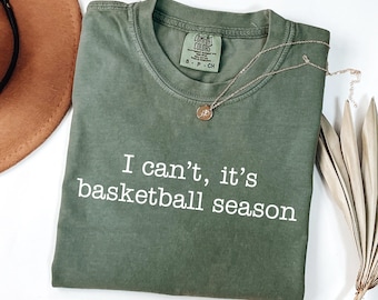 Pode incluir: Uma t-shirt de manga curta verde-azeitona com gola redonda e o texto branco "I can't, it's basketball season" impresso na frente. A t-shirt apresenta uma etiqueta da marca Comfort Colors no interior da gola e um pequeno colar com pendente dourado.