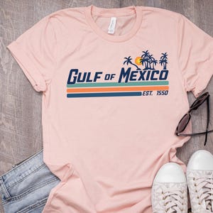May include: A pink t-shirt with a retro-style graphic that says "GULF of MEXICO EST. 1950" with palm trees and a sun.