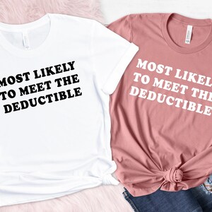 May include: Two t-shirts, one white and one dusty rose, with the text "MOST LIKELY TO MEET THE DEDUCTIBLE" in bold, black and white font. The white shirt is on the left, and the dusty rose shirt is on the right, with a pair of blue jeans.