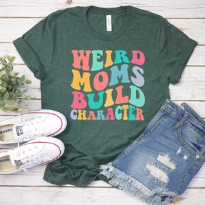Pode incluir: Uma camiseta verde com o texto "Weird Moms Build Character" em fonte retr&ocirc; colorida.